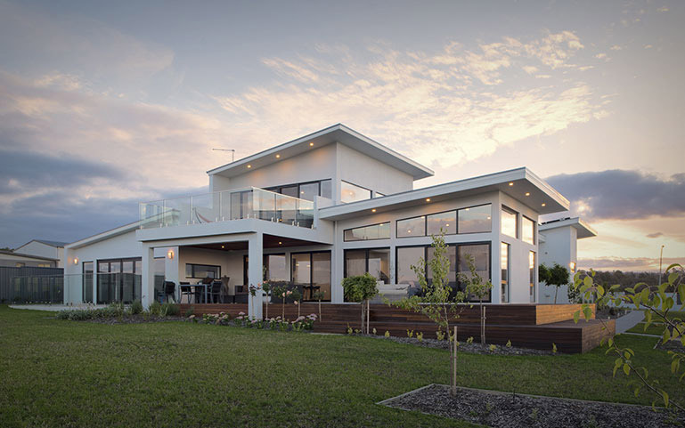 A beautiful white rendered two storey home photographed at dusk with a gorgeous modern style balcony and below a timber deck area for entertaining. 