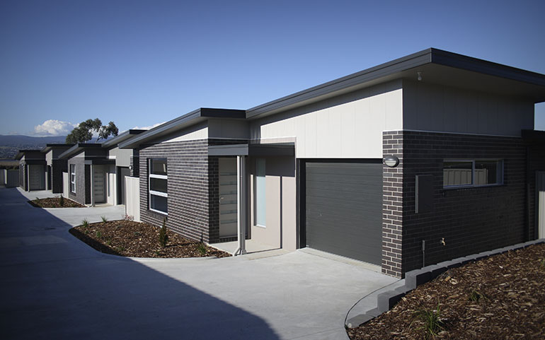 A row of compact identical brick units with slanting skillion roofs.  Each has a charcoal single garage and is accented with cream for the front door and surrounding the garage. 