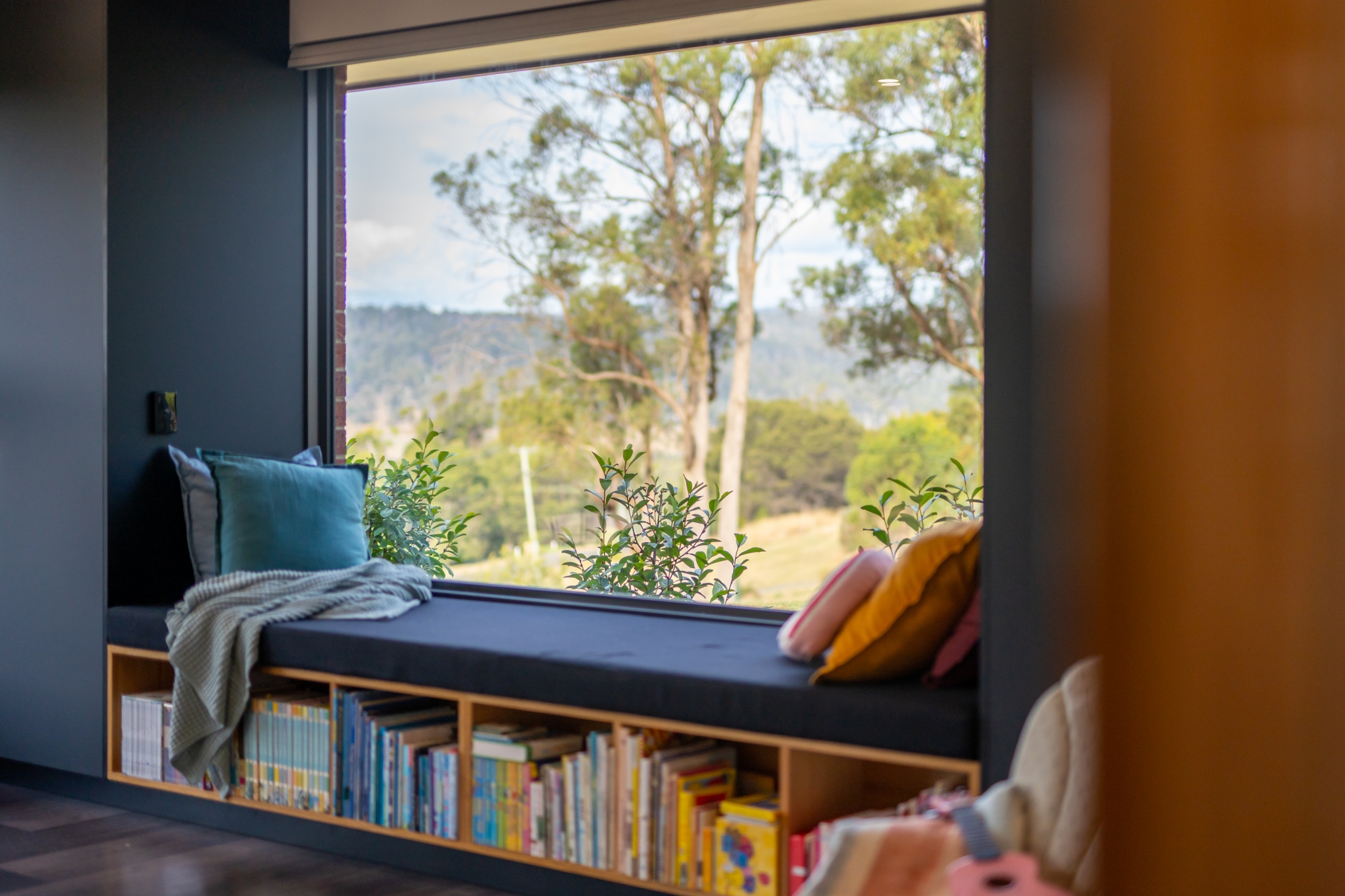 A window seat with storage for childrens books below and a window looking out to a bushy landscape